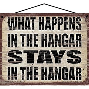 May include: A rustic wooden sign with distressed white paint and black lettering. The sign reads "What Happens In The Hangar Stays In The Hangar".