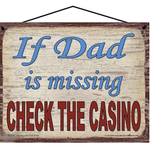 May include: A rustic wooden sign with a distressed beige background and brown edges. The sign has the text "If Dad is missing CHECK THE CASINO" in blue and red lettering. The sign also includes the text "Hip Signs for Cool Folks" and "Lorrie Powell Studios Made in USA".
