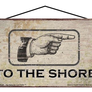 May include: A rustic wooden sign with a distressed white background and black text that reads "TO THE SHORE". A black and white illustration of a pointing hand is above the text. The sign has a distressed look and is meant to be hung.