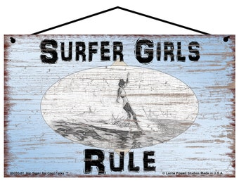 May include: A blue and white wooden sign with the text "Surfer Girls Rule" in black lettering. The sign features a black and white illustration of a surfer girl riding a wave.