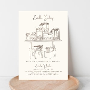 May include: An invitation card with a bakery theme, featuring a line drawing of baked goods displayed on a table. The text reads "Estelle's Bakery" and invites guests to celebrate the mom-to-be. Includes event details and RSVP information.