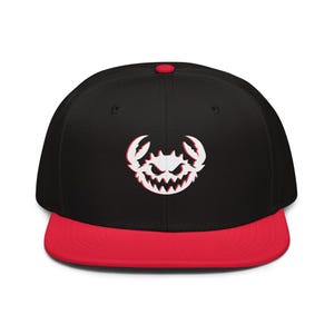Könnte beinhalten: Eine schwarze Snapback-Cap mit flachem rotem Schirm und einem roten Knopf auf der Oberseite. Auf der Vorderseite befindet sich eine weiße Grafik eines stilisierten, aggressiven Krebses mit spitzen Zähnen und Scheren, betont durch einen roten Schatten.