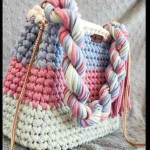 Puede incluir: Bolso de cubo hecho a mano en crochet en colores pastel. El bolso presenta un asa trenzada en rosa, azul y blanco, con una borla a juego. El cuerpo del bolso es una mezcla de puntadas de ganchillo rosas, azules y blancas, con una correa de cadena dorada.