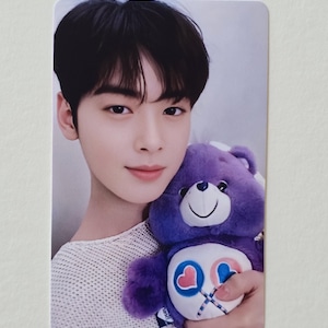 May include: A photograph of a person holding a purple Care Bear plush toy. The person has dark hair and is wearing a white knit top. The Care Bear has a heart and lollipop design on its belly.