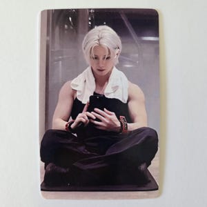 May include: A person with white hair wearing a black tank top and black pants, sitting cross-legged and holding a black phone. A white towel is around the neck. The person is wearing a red watch.