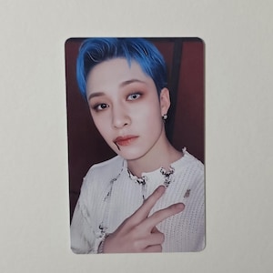 May include: A rectangular trading card showing a person with blue hair and a white sweater. The person is making a peace sign with their fingers. The background is a dark red colour.