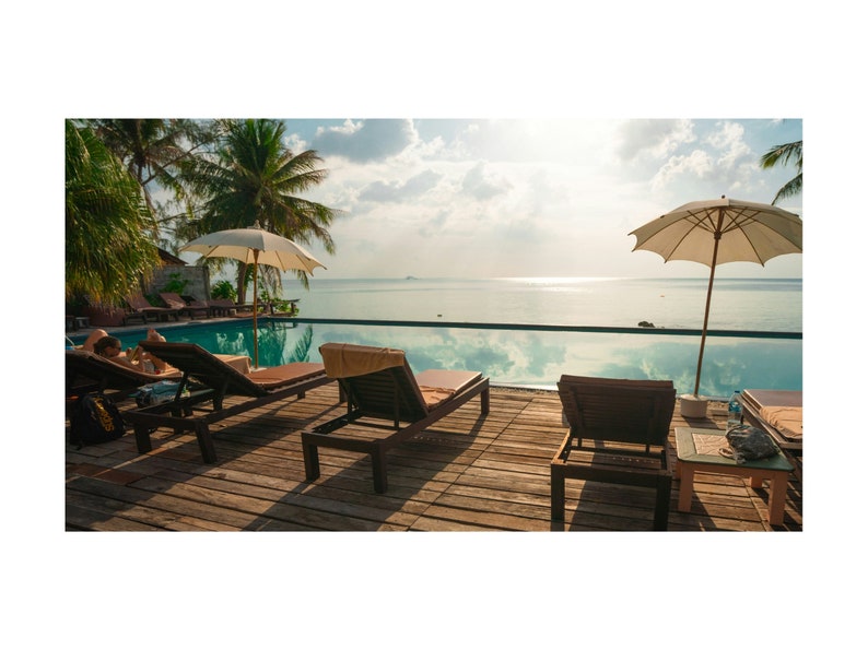 May include: A swimming pool with lounge chairs and umbrellas on a wooden deck overlooking the ocean. The pool is surrounded by lush tropical greenery.