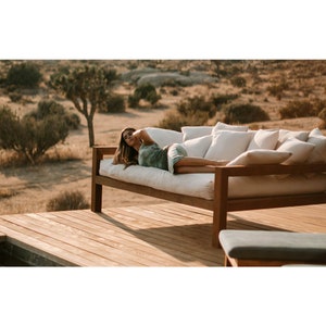 May include: A woman relaxes on a wooden outdoor daybed with white cushions. The daybed is on a wooden deck overlooking a desert landscape.