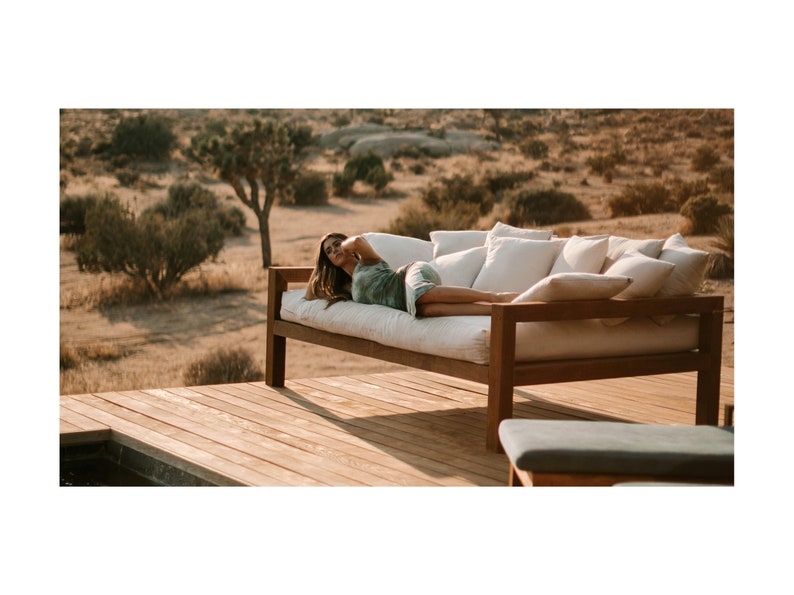 May include: A woman relaxes on a wooden outdoor daybed with white cushions. The daybed is on a wooden deck overlooking a desert landscape.