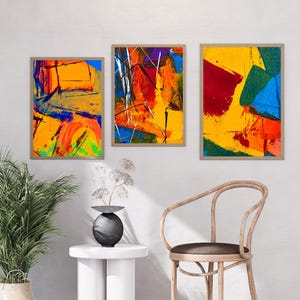 Puede incluir: Tres impresiones artísticas abstractas enmarcadas con colores vibrantes. La obra presenta pinceladas audaces en tonos de amarillo, naranja, rojo, azul y verde. Los marcos son de color marrón neutro y el arte se exhibe en una pared blanca.