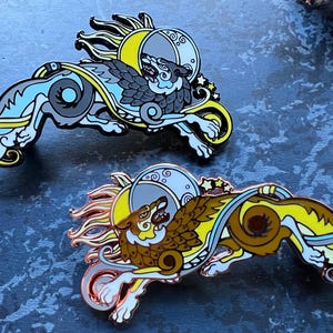 May include: Two enamel pins featuring a wolf design with a sun and moon motif. One pin has a blue, gray, and yellow color scheme, while the other is brown, yellow, and copper. The pins have intricate details and a stylized aesthetic.