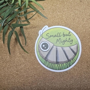Może przedstawiać: Okrągła naklejka z zielono-białą obwódką, przedstawiająca kreskówkowy wzór z dużym okiem i segmentowym korpusem. Tekst "Small but Mighty" jest napisany czcionką skryptową.