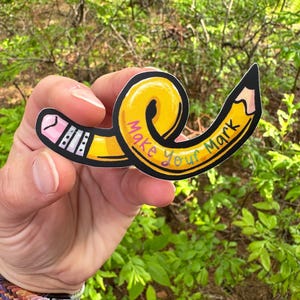 May include: A colorful sticker in the shape of a yellow pencil with the text "Make Your Mark". The pencil has a pink eraser and a black tip. The sticker is held in front of a green leafy background.