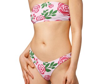 May include: A two-piece bikini set featuring a floral pattern of pink roses and green leaves on a light pink background. The strapless top and low-rise bottoms are made of a soft, likely stretchy material.
