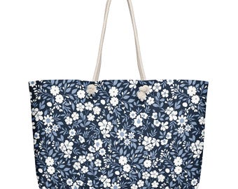 May include: A large tote bag with a floral pattern. The bag features a navy blue background covered in white and light blue flowers and leaves. It has two cream-colored rope handles.