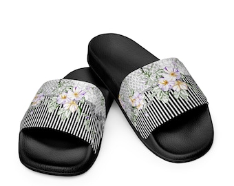 May include: Black slide sandals with a floral design. The sandals feature a black and white striped pattern on the front, with a floral print in white, purple, and green. The sandals are made of a rubber-like material.