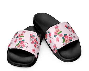 May include: Black slide sandals with a light pink strap featuring a floral pattern of pink and red flowers with green leaves. The sandals are angled towards the viewer, showcasing the design.