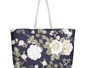 May include: A large tote bag with a floral pattern. The bag features a navy blue background with white roses and green leaves. The handles are made of a thick, off-white rope.