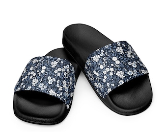May include: A pair of black slide sandals with a floral pattern. The top of each sandal features a navy blue background with a repeating pattern of small white flowers and blue leaves. The sandals are set against a white background.
