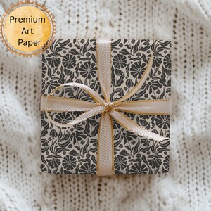 Puede incluir: Una caja de regalo cuadrada envuelta en papel con estampado floral, atada con una cinta dorada. El papel presenta un dise&ntilde;o repetido de flores y hojas de color gris oscuro sobre un fondo crema. Las palabras "Premium Art Paper" est&aacute;n en un gr&aacute;fico circular.