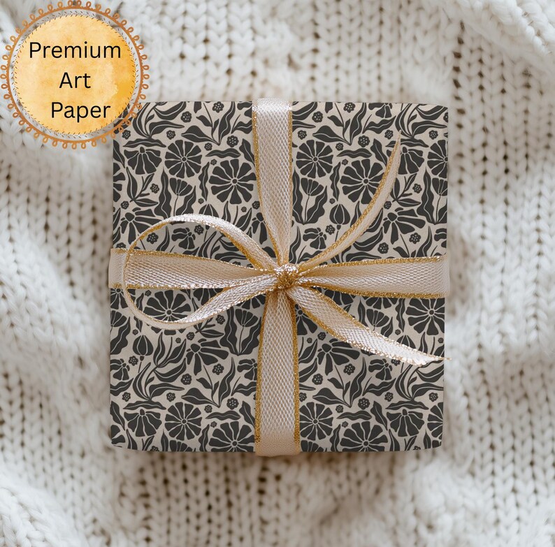 Puede incluir: Una caja de regalo cuadrada envuelta en papel con estampado floral, atada con una cinta dorada. El papel presenta un dise&ntilde;o repetido de flores y hojas de color gris oscuro sobre un fondo crema. Las palabras "Premium Art Paper" est&aacute;n en un gr&aacute;fico circular.