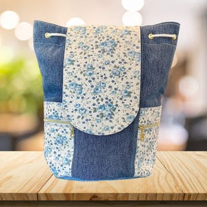 May include: A denim backpack with a floral flap and side pockets. The flap and pocket accents feature a white fabric with blue floral patterns. The backpack has a drawstring closure and gold-colored zippers.