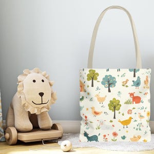 May include: A cream-colored tote bag with a whimsical farm animal and tree pattern. The bag has a beige handle and is next to a wooden lion toy. The design includes birds, trees, and a barn.