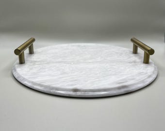 Puede incluir: Una bandeja redonda de mármol con asas doradas. La bandeja es blanca con vetas grises y tiene dos asas doradas en forma de barra en lados opuestos. Diseñada para servir alimentos o exhibir objetos.