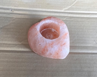 May include: A heart-shaped, pink Himalayan salt candle holder. The candle holder has a circular indentation in the center for a tea light. The salt has a natural, textured appearance with variations in color.