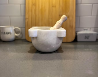 May include: A white marble mortar and pestle set with a wooden pestle. The mortar has a rounded base and two handles. A light brown wooden cutting board is in the background. A white mug with the word "escape" is on the left.