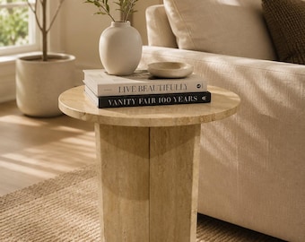 May include: A round beige travertine side table with a cross-shaped base. On the table surface sits a white ceramic vase containing green leafy branches, a small beige bowl, and two stacked books titled Live Beautifully and Vanity Fair 100 Years.