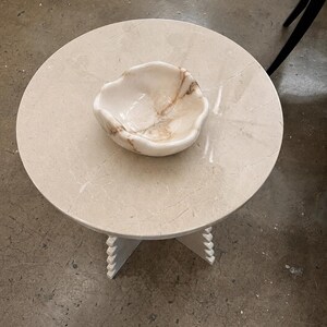 May include: A round beige stone side table with a smooth surface and a cross-shaped base featuring notched edges. Centered on the table is a small, organic-shaped bowl made of polished white and tan marble with visible natural veining throughout.