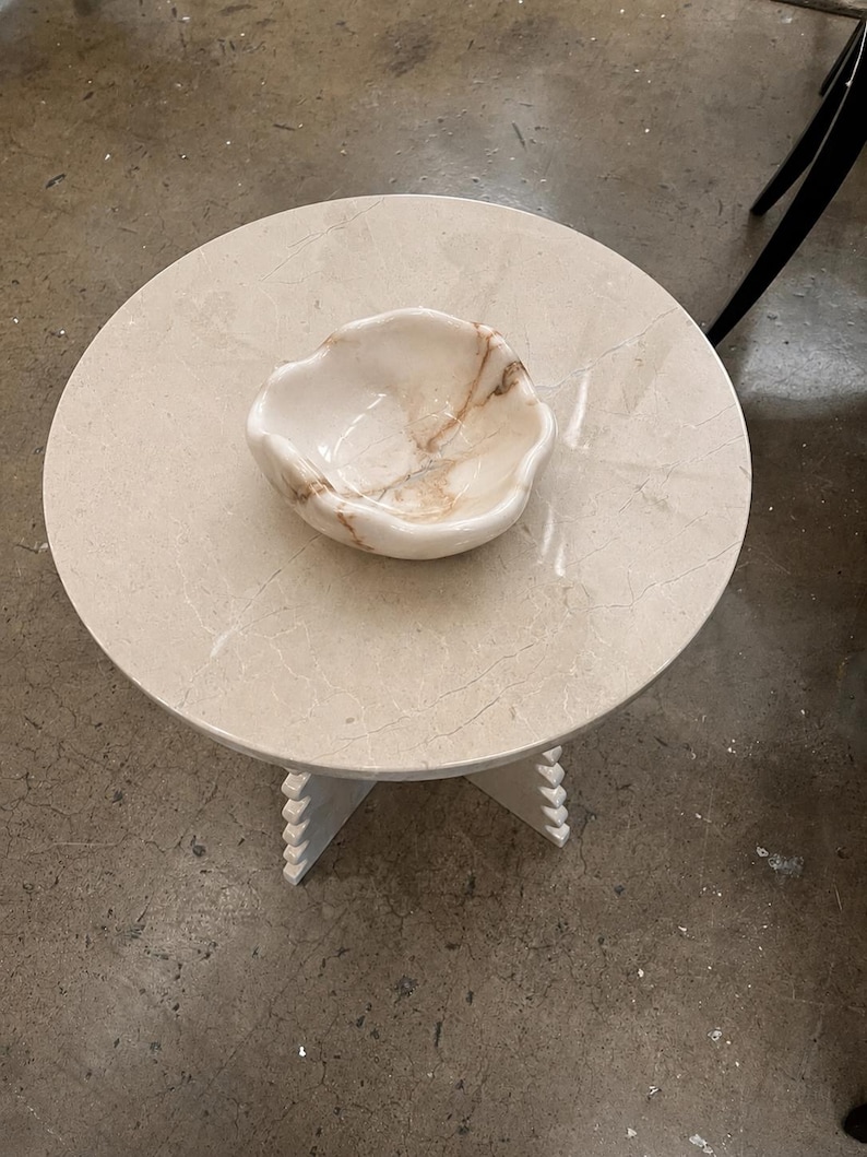 May include: A round beige stone side table with a smooth surface and a cross-shaped base featuring notched edges. Centered on the table is a small, organic-shaped bowl made of polished white and tan marble with visible natural veining throughout.