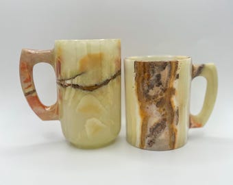 May include: Two onyx mugs with unique patterns. The larger mug has a handle with orange and brown hues, while the smaller mug features a handle with a similar color scheme. Both mugs have a light green and brown color palette.