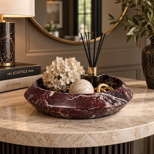May include: A circular bowl crafted from deep red marble with prominent white veining. The bowl contains a cluster of cream-colored hydrangea flowers, a smooth beige stone sphere, a glass reed diffuser with black sticks, and a gold-toned metal chain link.