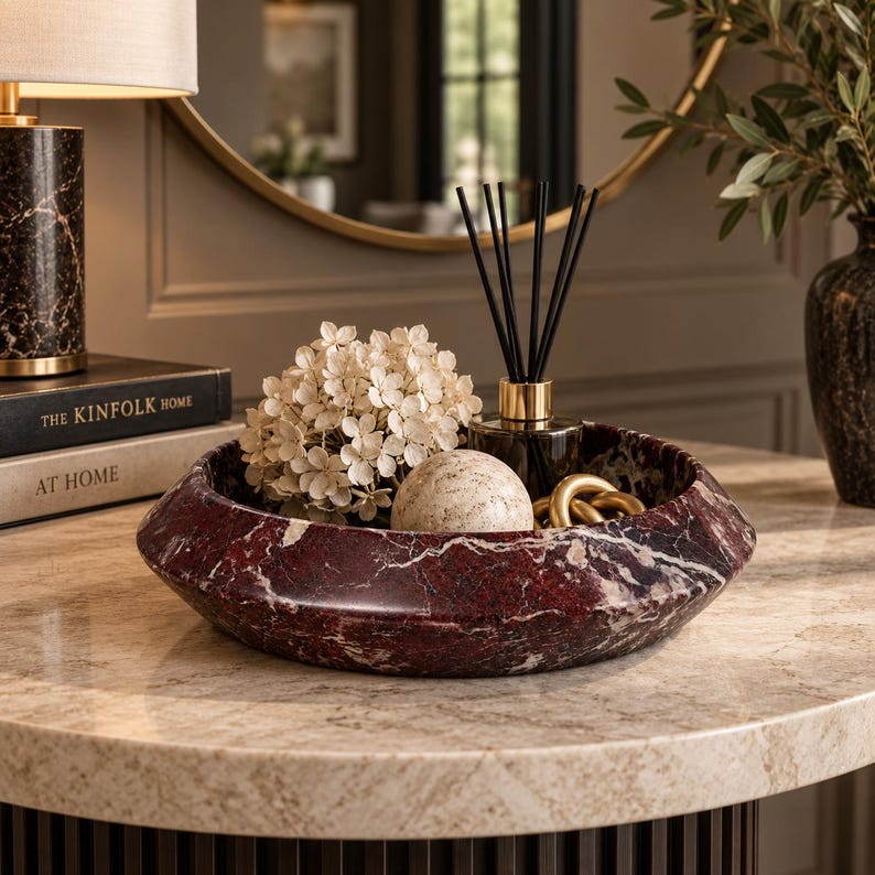 May include: A circular bowl crafted from deep red marble with prominent white veining. The bowl contains a cluster of cream-colored hydrangea flowers, a smooth beige stone sphere, a glass reed diffuser with black sticks, and a gold-toned metal chain link.
