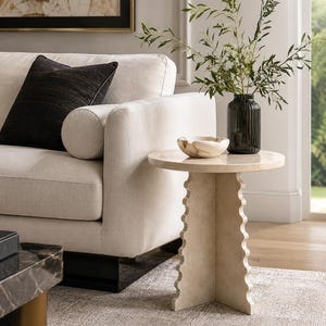 May include: A round side table with a smooth top and a base featuring three vertical panels with scalloped edges. The table is crafted from a light beige stone material. A small, light-colored stone bowl and a dark green ribbed glass vase are placed on top.