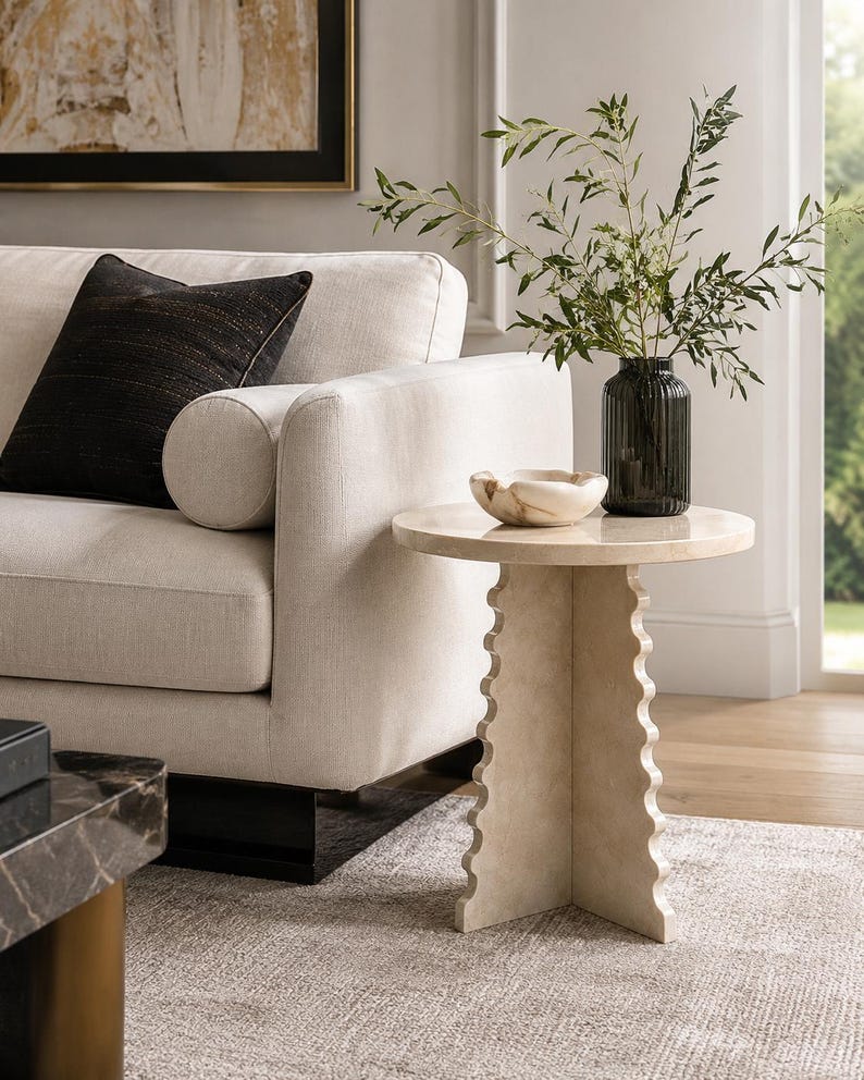 May include: A round side table with a smooth top and a base featuring three vertical panels with scalloped edges. The table is crafted from a light beige stone material. A small, light-colored stone bowl and a dark green ribbed glass vase are placed on top.