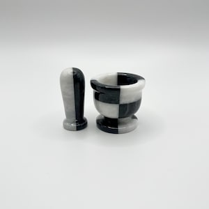 May include: A black and white marble mortar and pestle set. The mortar is a small bowl with a checkered pattern, and the pestle is a rounded, cylindrical shape. The set is designed for grinding herbs and spices.