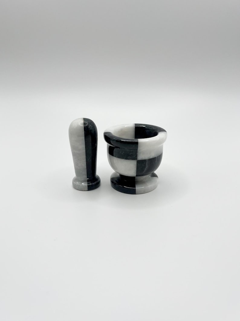 May include: A black and white marble mortar and pestle set. The mortar is a small bowl with a checkered pattern, and the pestle is a rounded, cylindrical shape. The set is designed for grinding herbs and spices.