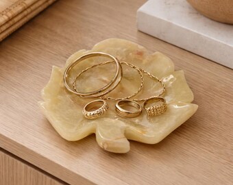 Puede incluir: Una bandeja de joyería de ónix amarillo claro en forma de hoja contiene varios anillos y pulseras doradas. La bandeja se encuentra sobre una superficie de madera clara, con una caja de madera y un cuenco de cerámica en el fondo. La joyería incluye una variedad de estilos de anillos.