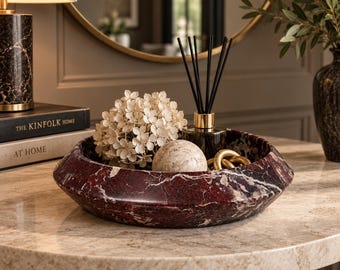 May include: A circular bowl crafted from deep red marble with prominent white veining. The bowl contains a cluster of cream-colored hydrangea flowers, a smooth beige stone sphere, a glass reed diffuser with black sticks, and a gold-toned metal chain link.