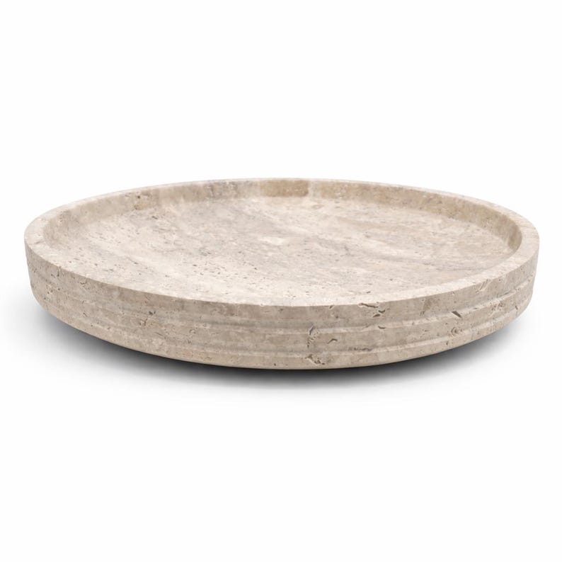 Puede incluir: Una bandeja circular de piedra beige con una textura natural porosa y un borde elevado. La bandeja presenta una paleta de colores bronceado claro con variaciones sutiles en la veta de la piedra, una superficie interior lisa y un borde exterior.