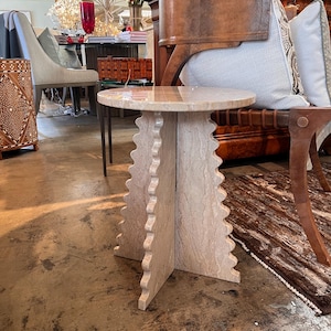 Pode incluir: Uma mesa lateral de pedra bege com um tampo redondo e uma base &uacute;nica com bordas onduladas. A mesa &eacute; feita de pedra de cor clara e tem uma apar&ecirc;ncia natural e texturizada. A mesa tem aproximadamente 50 cm de di&acirc;metro e 50 cm de altura.