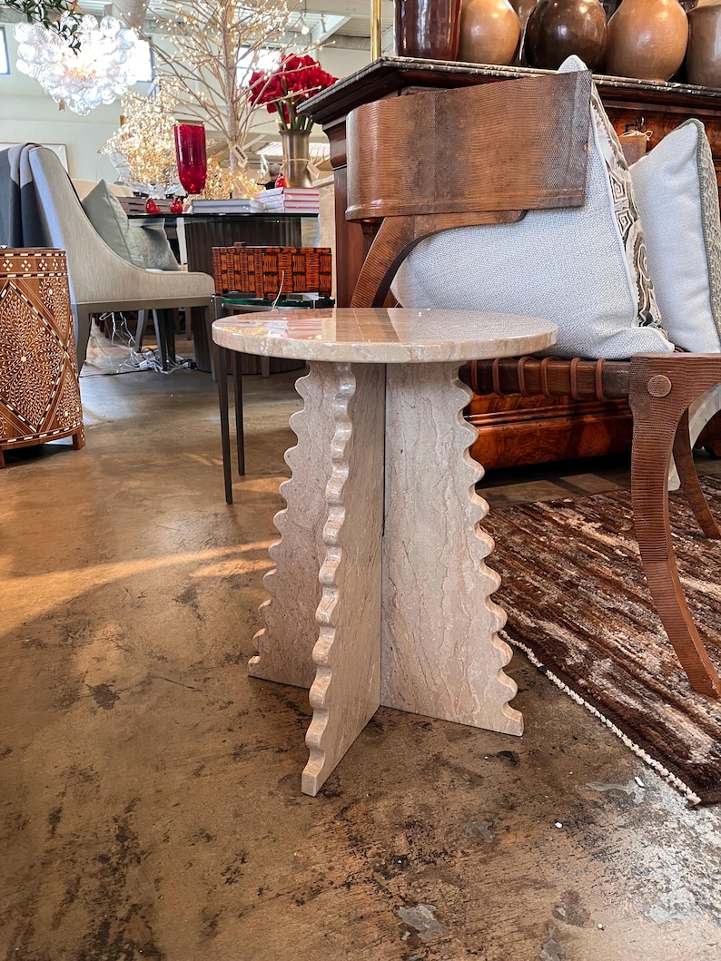 Pode incluir: Uma mesa lateral de pedra bege com um tampo redondo e uma base &uacute;nica com bordas onduladas. A mesa &eacute; feita de pedra de cor clara e tem uma apar&ecirc;ncia natural e texturizada. A mesa tem aproximadamente 50 cm de di&acirc;metro e 50 cm de altura.