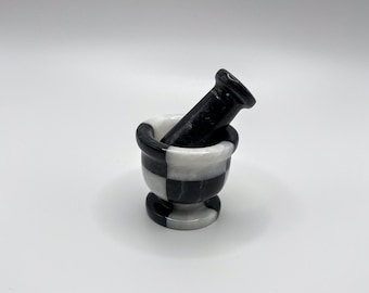 May include: A small, decorative mortar and pestle set. The mortar is a bowl-shaped vessel with a checkered black and white pattern. The pestle is a solid black, cylindrical tool. The set is made of stone and is suitable for grinding herbs and spices.