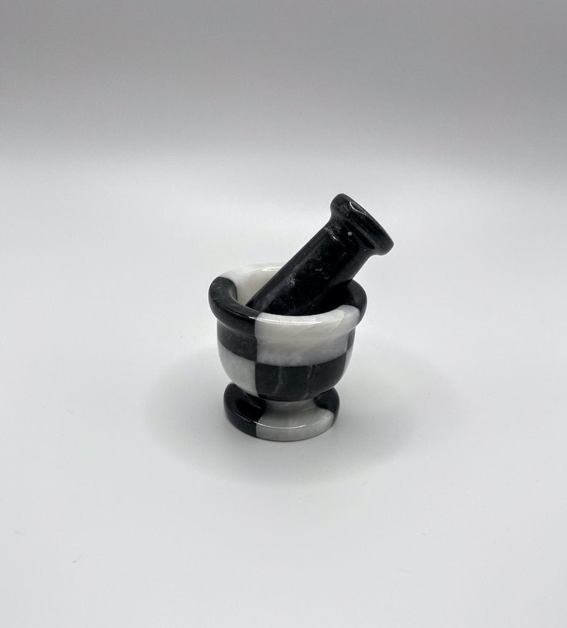 May include: A small, decorative mortar and pestle set. The mortar is a bowl-shaped vessel with a checkered black and white pattern. The pestle is a solid black, cylindrical tool. The set is made of stone and is suitable for grinding herbs and spices.