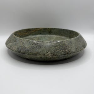 May include: A circular stone bowl featuring a mottled green and grey surface with natural veining. The bowl has a wide, sloped rim that tapers towards the base, creating a low-profile, heavy appearance with a smooth, polished finish throughout.
