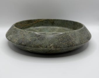 Puede incluir: Un cuenco circular de piedra con una superficie moteada en tonos verdes y grises con vetas naturales. El cuenco tiene un borde ancho e inclinado que se estrecha hacia la base, creando una apariencia baja y pesada con un acabado pulido.