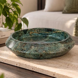 May include: A circular, shallow bowl crafted from polished green stone with natural dark veining and a smooth, glossy finish. The bowl features a wide, sloped rim and a deep center, showcasing the unique mineral patterns and textures of the stone.